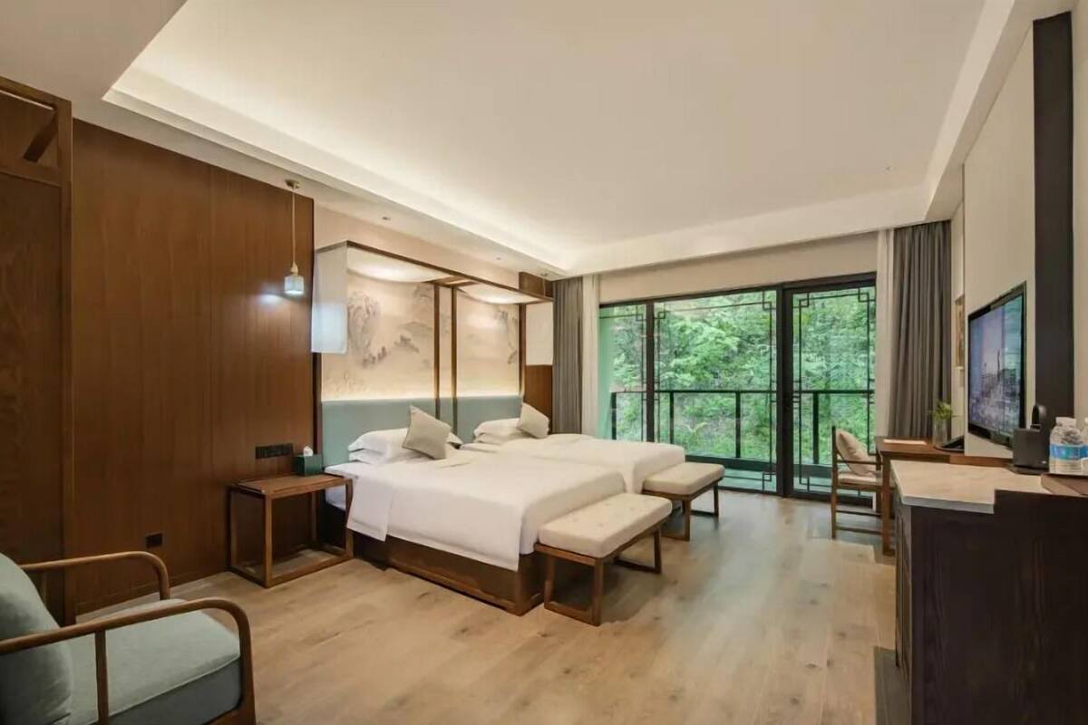 A modern hotel room with a large bed, a desk, and a view of greenery through the windows.