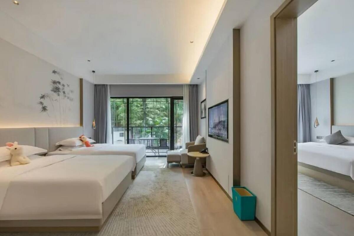 A modern hotel room with two beds, a balcony with a view of greenery, a small seating area, and a painting on the wall.