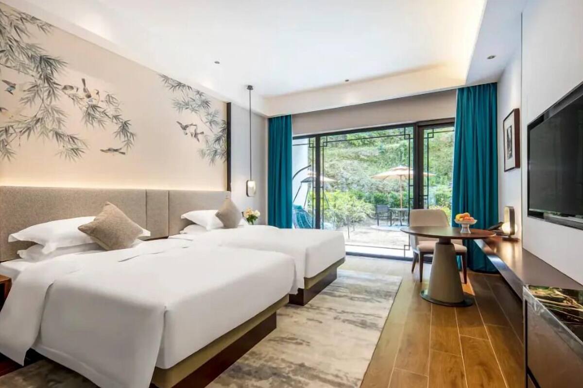 A modern hotel room with two beds, a dining area, a TV, and a balcony with a view of greenery.
