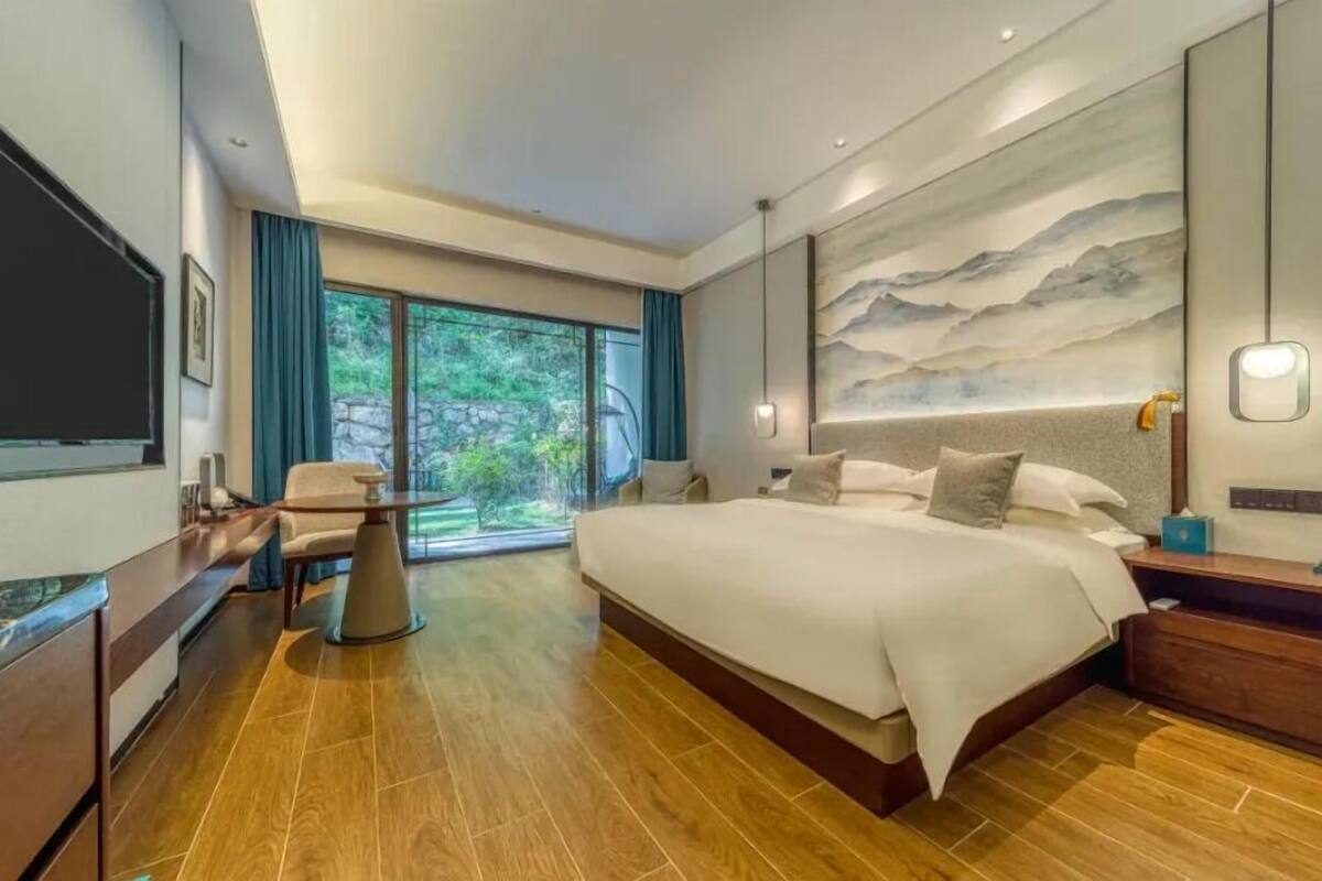 A modern hotel room with a large bed, a desk, and a view of greenery through the window.
