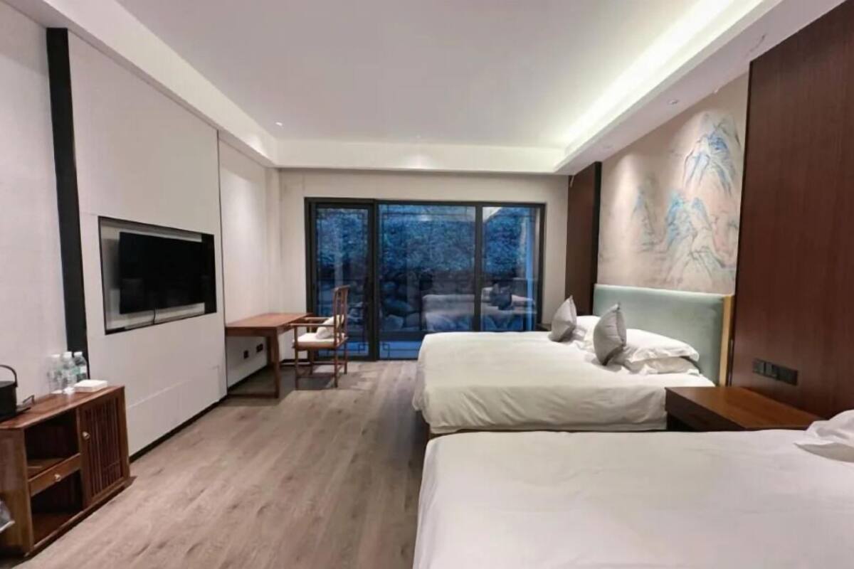 A modern hotel room with two beds, a desk, a television, and a sliding glass door to the outside.