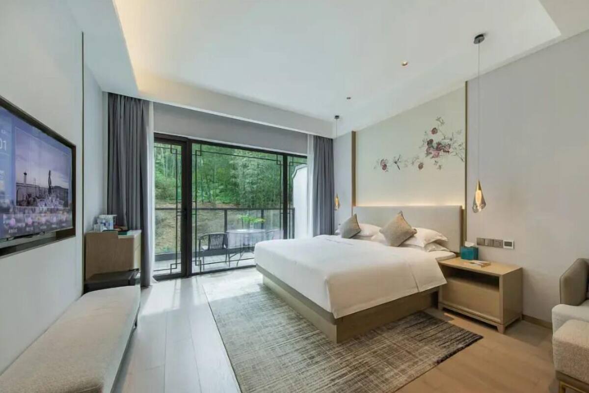 A modern bedroom with a large bed, a nightstand, a sofa, and a balcony with a view of greenery.