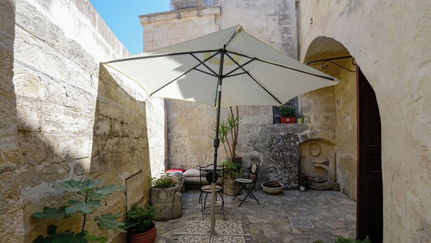 A stone courtyard with a table and chairs under a large umbrella, surrounded by potted plants and a statue. - Lovely house with AC in Matera - Perfect for a getaway (Matera)