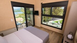 A bedroom with a bed, a sliding glass door, a TV, and a view of a lake and greenery. - VILLA PENELOPE
 (Katouna)