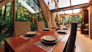 A dining area with a long wooden table, woven chairs, and a view of lush greenery through large windows. - Spectacular Dune Villa with a private sauna and breathtaking views of the dunes (Soest)