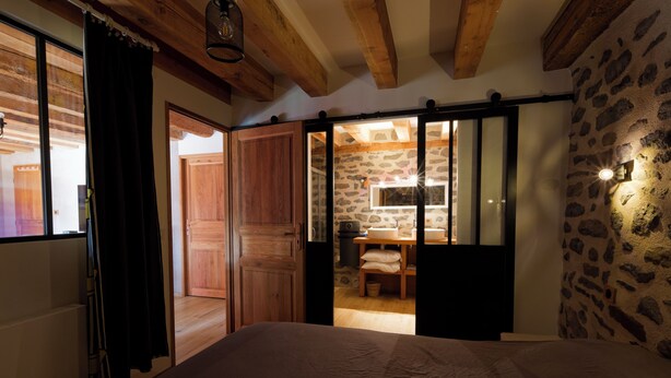 Un dormitorio con una cama grande, vigas de madera en el techo, una pared de piedra y un acceso corredizo a una zona de cocina.