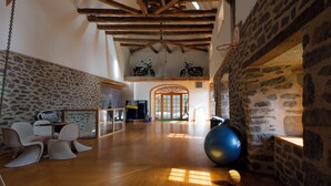 Una habitación amplia con techo alto, paredes de piedra y vigas de madera. Se observa una pelota grande para ejercicios, un aro de baloncesto y un área de descanso con sillas.