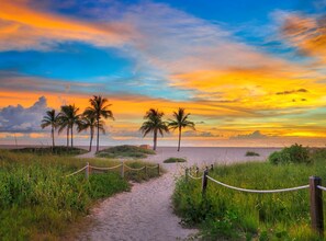 Ein Weg führt zu einem Strand mit Palmen und Blick auf den Sonnenuntergang.