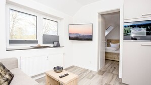 Una habitación moderna y luminosa con un amplio ventanal, un televisor de pantalla plana, una pequeña mesa de madera y un rincón de descanso acogedor.