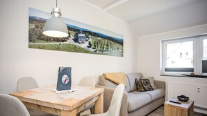 Una sala de estar moderna con una mesa de comedor de madera, dos sillas grises, un sofá beige y un taburete de madera pequeño. Un cuadro de paisaje grande decora la pared, y una ventana proporciona luz natural.