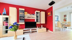 A modern living room with a red accent wall, a flat-screen TV, a white coffee table, and a bookshelf. - Fully equipped vacation apartment with plenty of space, barbecue and garden, centrally located (Waltershausen)