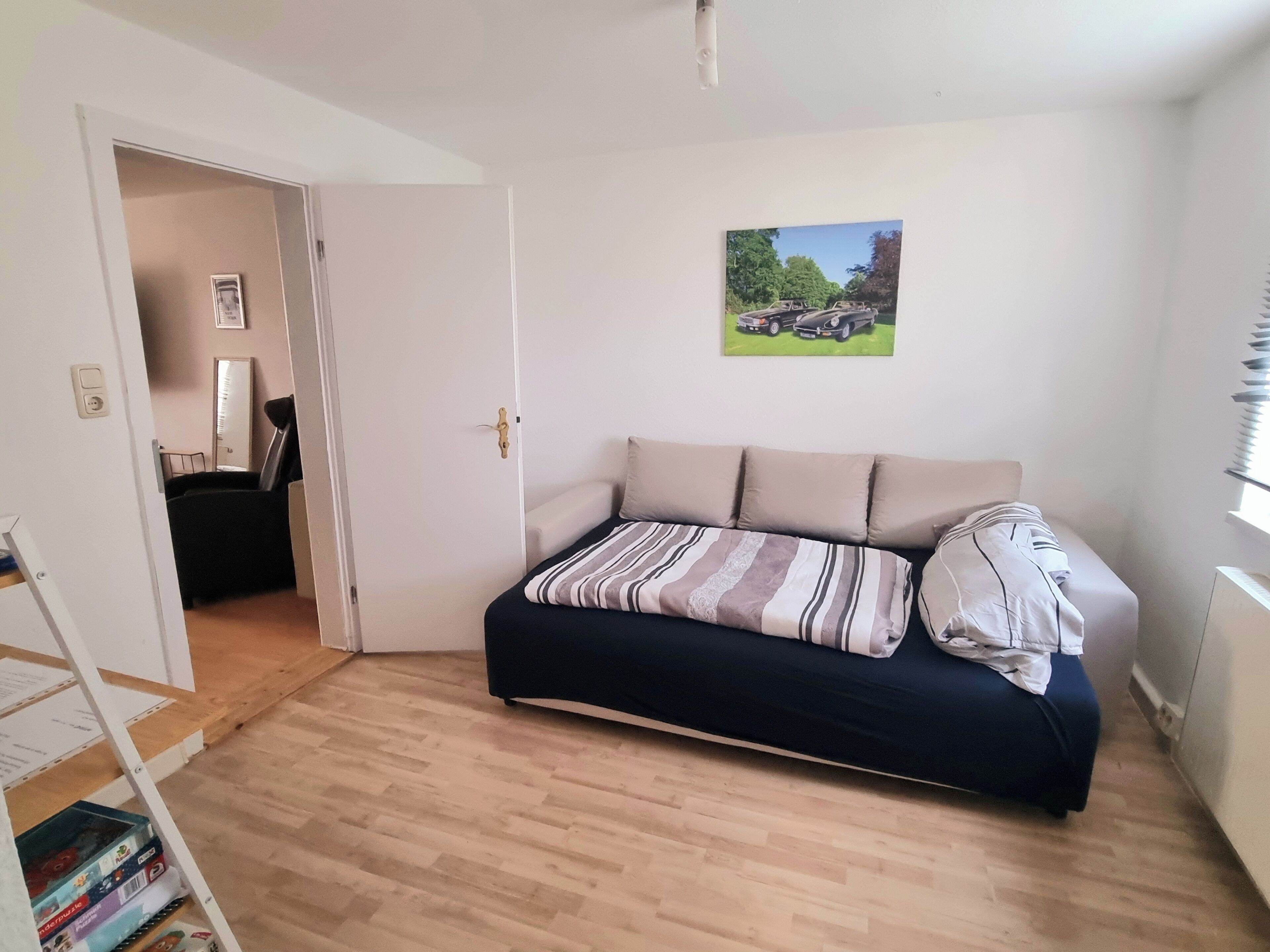 A room with a bed, a door leading to another room, a small table, and a wall-mounted picture.