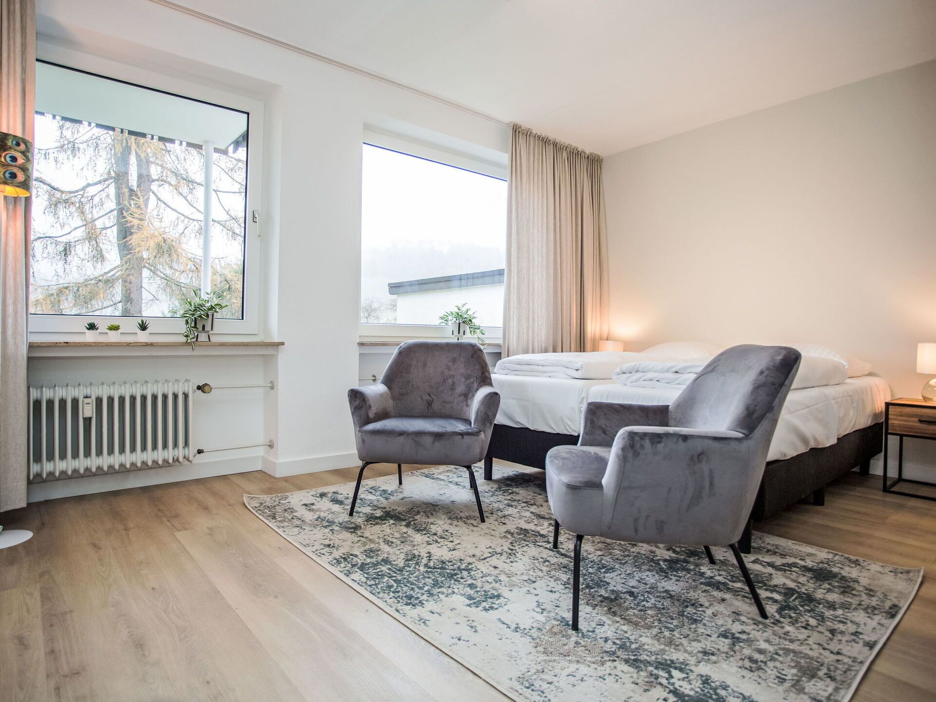 Quarto de hotel moderno com uma cama grande, duas poltronas, um radiador e vista para o exterior.