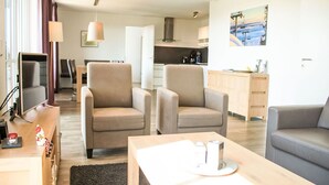 Un moderno salón con dos sillones beige, un sofá gris, una mesa de centro de madera y un televisor sobre un mueble de madera. Se aprecia una cocina al fondo, con un cuadro en la pared.