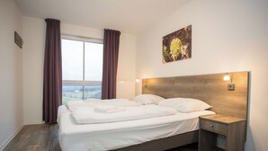 Habitación de hotel con una cama grande, una mesita de noche, una lámpara de pared y un ventanal con cortinas.