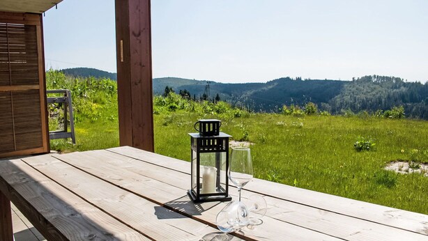 Una mesa de madera con un vaso, una vela, un farol, y vistas a un campo cubierto de hierba y colinas.