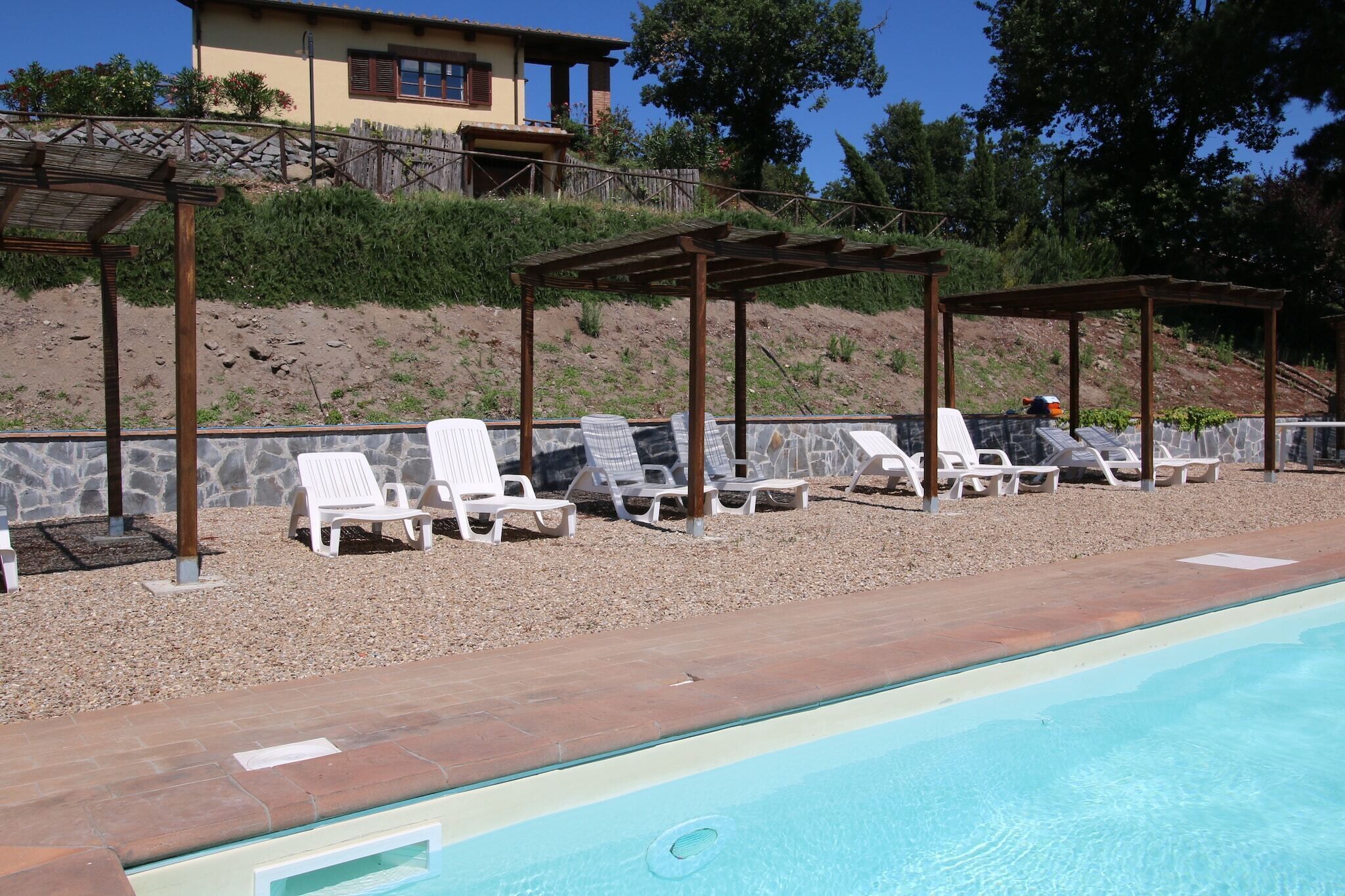 A poolside area with white lounge chairs under pergolas, a stone wall, and a building in the background.