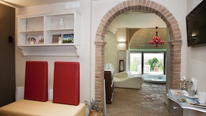 A room with a brick archway, a white shelf with decorative items, a red cushioned bench, a white sofa, and a television on the wall.
