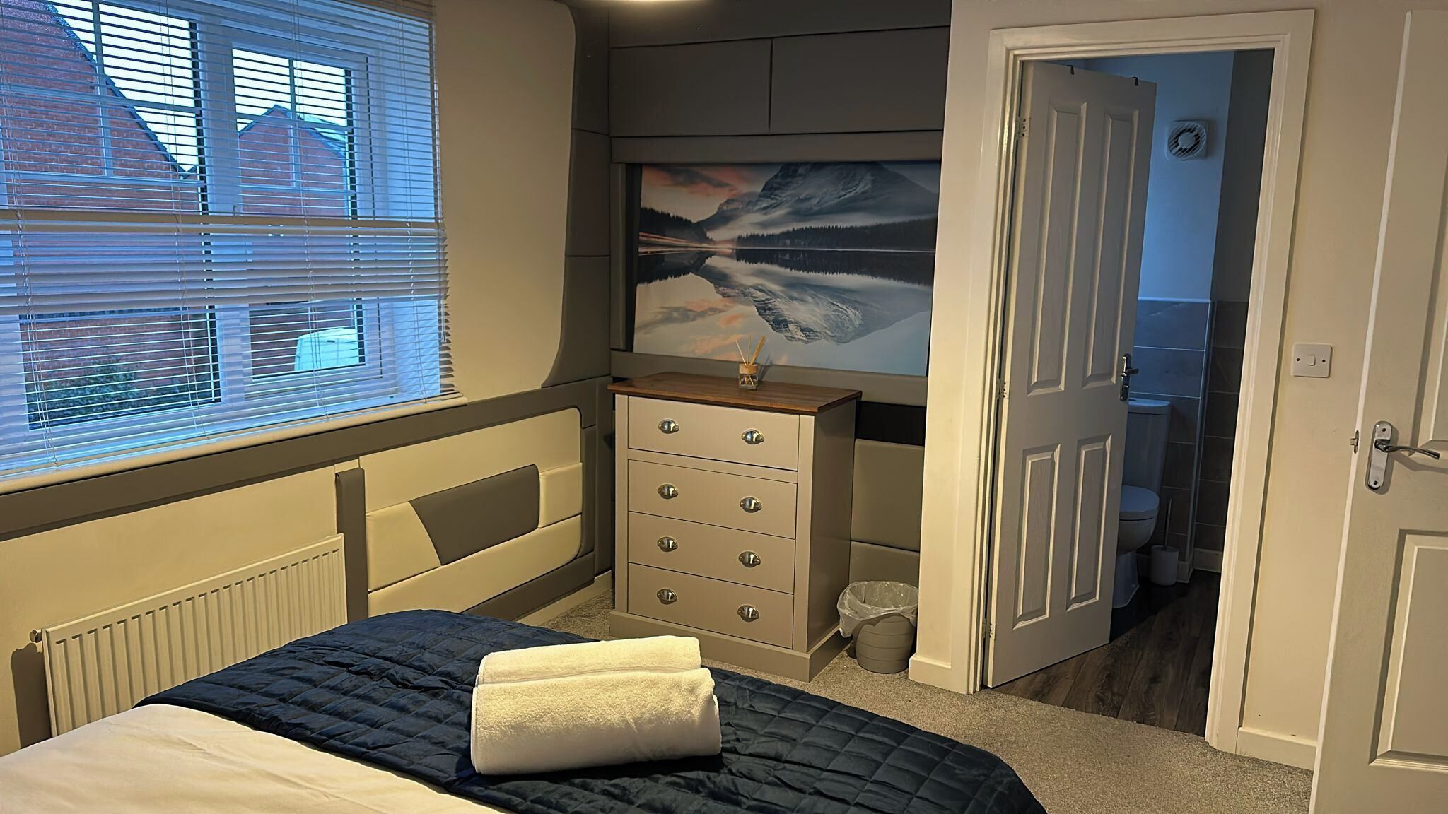 A bedroom with a bed, a dresser, a window with blinds, a painting on the wall, and a door leading to another room.