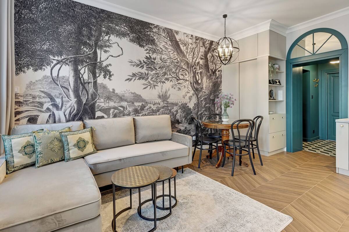 A living room with a large mural, a sofa with patterned cushions, a small round table, and a dining area with chairs.
