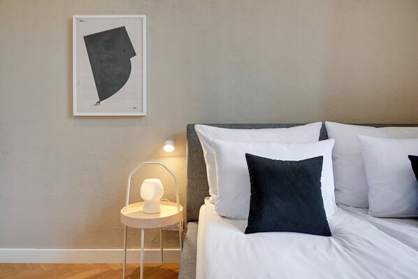 A bedroom with a bed, white and dark pillows, a bedside table with a lamp, and a framed abstract artwork on the wall.