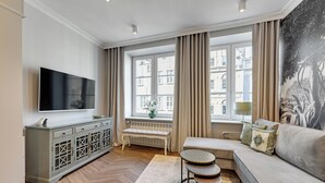 Een moderne woonkamer met een grote flatscreen televisie, een grijze kast met glazen deuren, een beige bank, een zitbank en een raam met gordijnen.