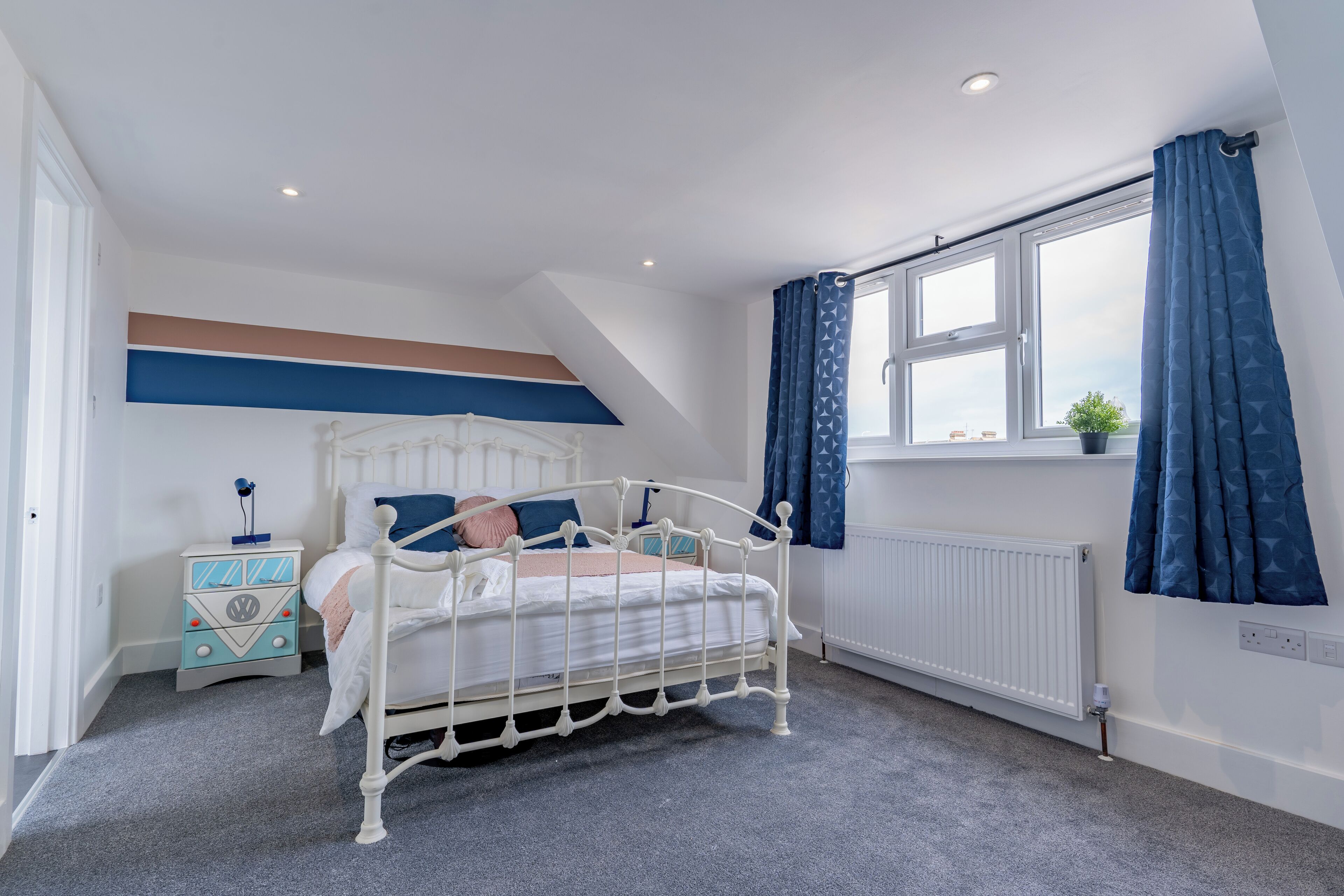 Une chambre à coucher avec un lit blanc, une table de chevet, une fenêtre avec des rideaux et un radiateur.