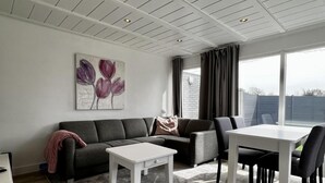 A modern living room with a grey sofa, a white coffee table, and a dining table with chairs. There is a painting on the wall and a window with curtains. - Beautiful Bungalow in North Holland (Sint Maartensvlotbrug)