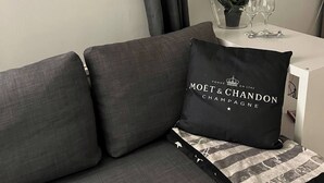 Een moderne woonkamer met een grijze bank, een zwart kussen met tekst, een hartvormige decoratie en een kleine tafel met een plant en glazen.