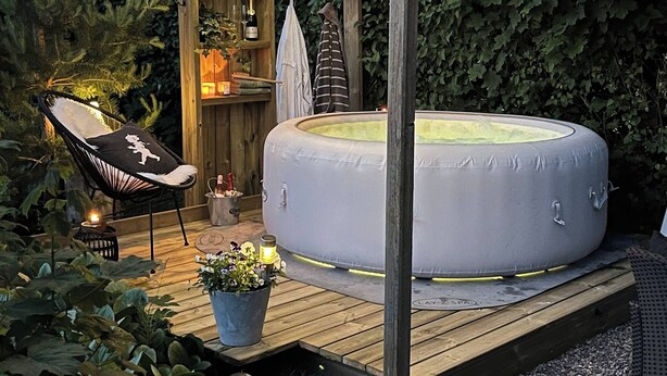 Een hottub op een houten terras, overdekt door een zonnescherm, omringd door groen en voorzien van een klein plankje met flessen en een handdoek.