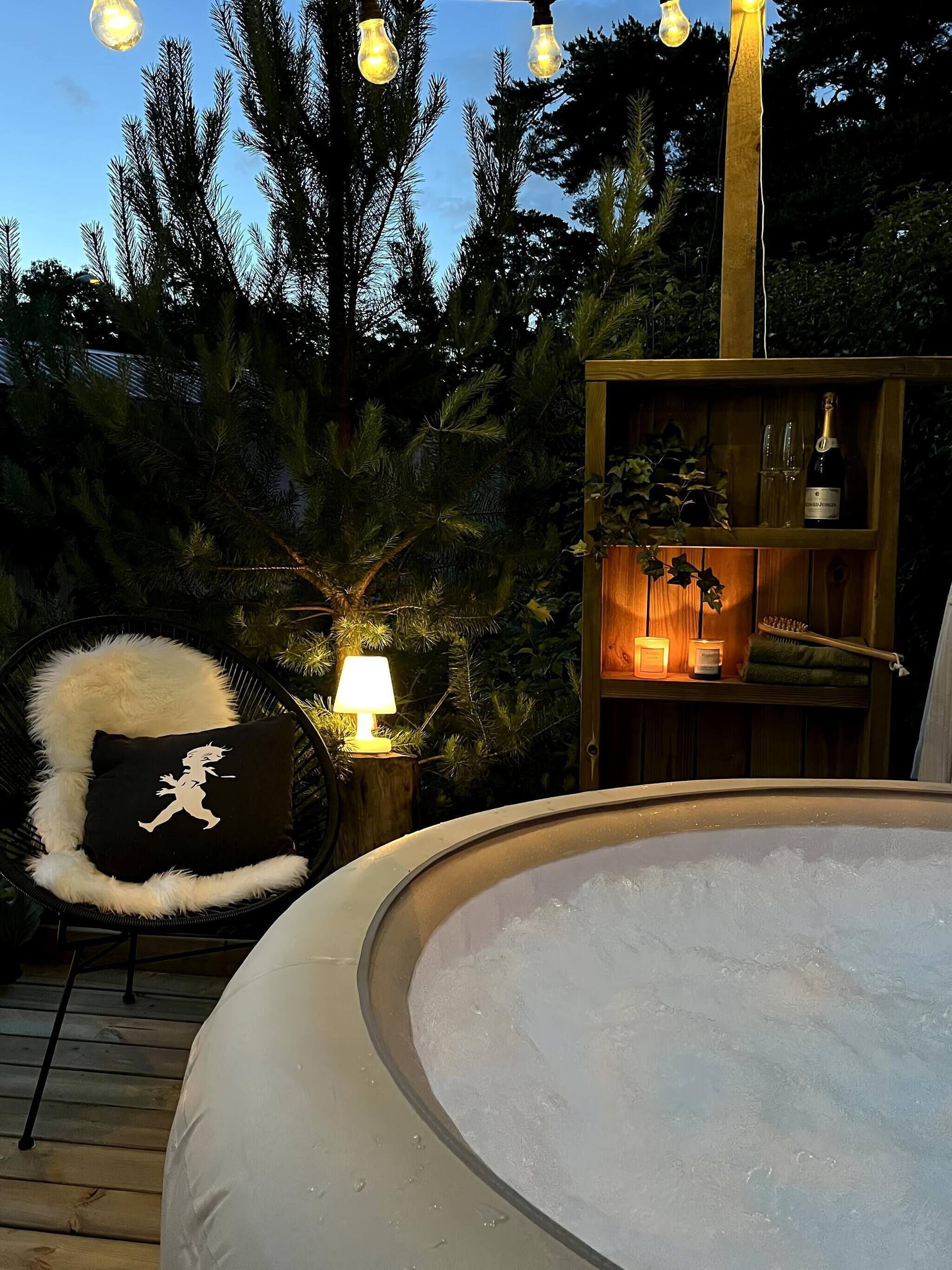 An outdoor hot tub surrounded by a wooden deck, with a black chair with a white cushion and a lamp nearby.