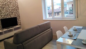 A living room with a sofa, a TV on a stand, a dining table with chairs, and a window with a view of buildings. - DOLCE NOTTE APARTMENTS (Verona)
