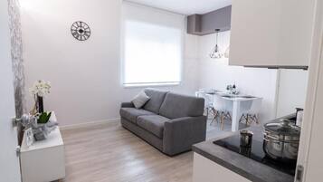 Een moderne woonkamer met een grijze bank, een klein wit bijzettafeltje met een vaas, een ronde wandklok en een raam met jaloezieën.