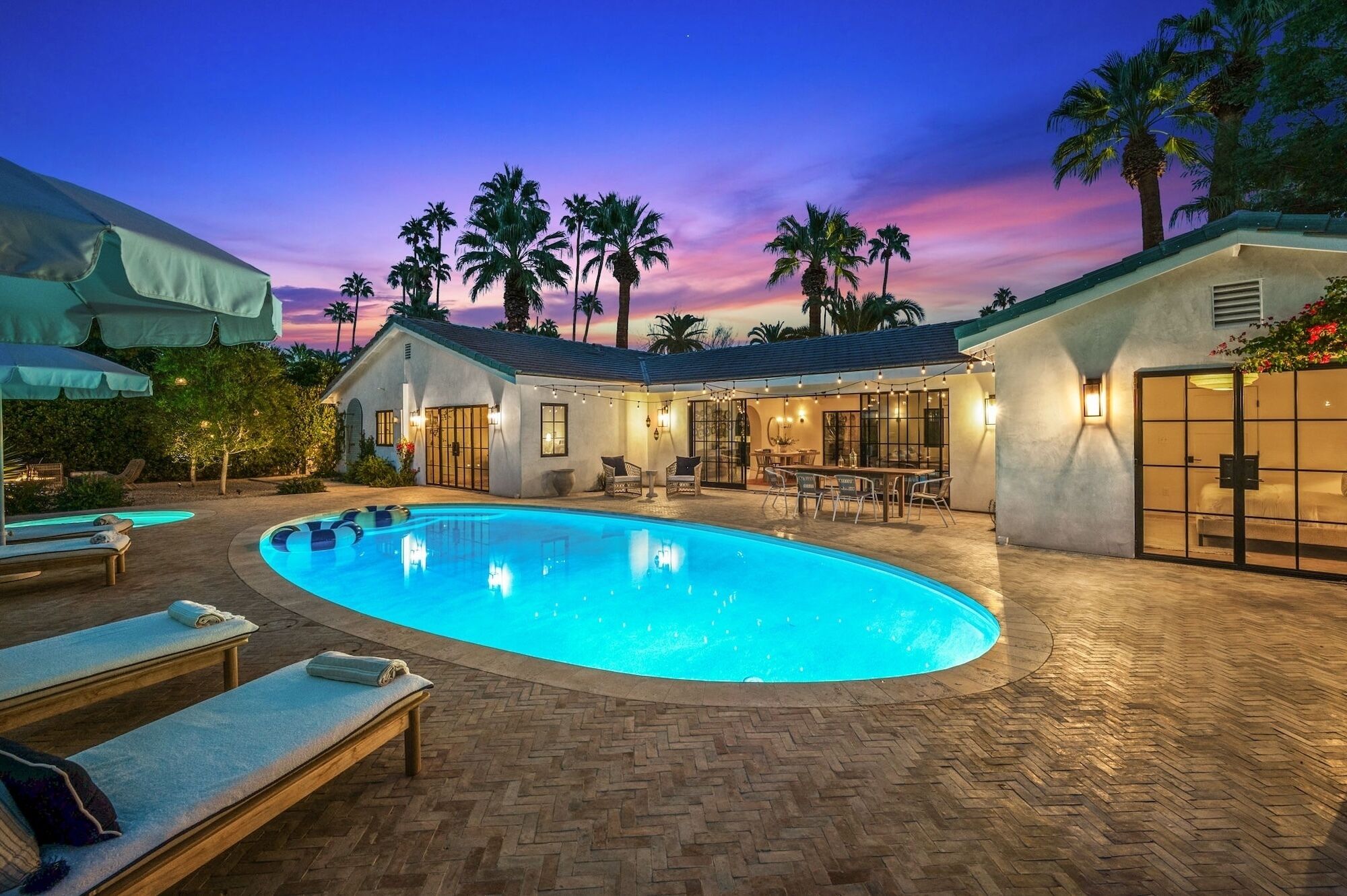 A poolside area with lounge chairs, a dining area, and a building with large windows and outdoor lighting.