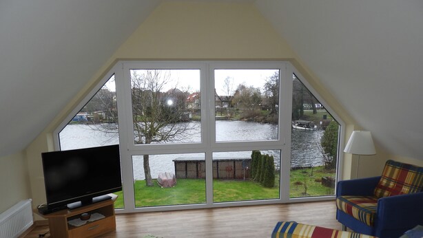 Ein Zimmer mit großem Fenster mit Blick auf ein Gewässer, einem Fernseher auf einem Holzboden und zwei Sesseln mit Karomuster.