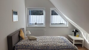 Ein Schlafzimmer mit einem Bett, zwei Fenstern, einem Heizkörper, einem Nachttisch und einer Lampe.