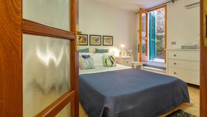 A bedroom with a bed, a nightstand, a dresser, and a window with a view of greenery. - Sardinia Re - Maiorca 13 (Alghero)