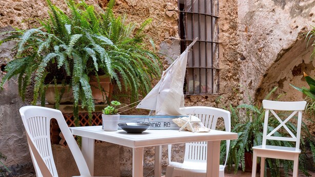 A white table and chairs set in a rustic outdoor area with terracotta tiles, surrounded by potted plants and a large fern. - Sardinia Re - Maiorca 13 (Alghero)