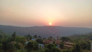 A rural landscape with a sunrise or sunset, houses, and a hilly terrain.