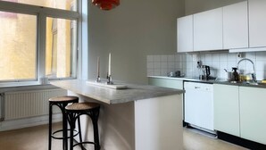 Een moderne keuken met een groot raam, witte kasten, een centraal eiland met krukken en een rode hanglamp.