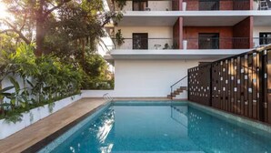 A swimming pool with a wooden deck, surrounded by greenery and a modern building with balconies. - Casa Aahana  by Limestays (Arpora)