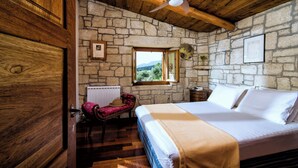 A bedroom with a stone wall, wooden ceiling beams, a bed with white linens, a wooden chair, and a window with a view. - Panoramic View Villa in OliveGroves (Loutraki)