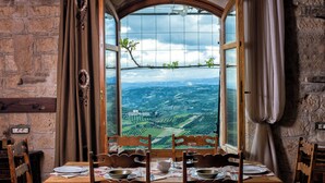 A dining table set with plates, glasses, and cutlery, a window with a view of a landscape, and a stone wall. - Panoramic View Villa in OliveGroves (Loutraki)