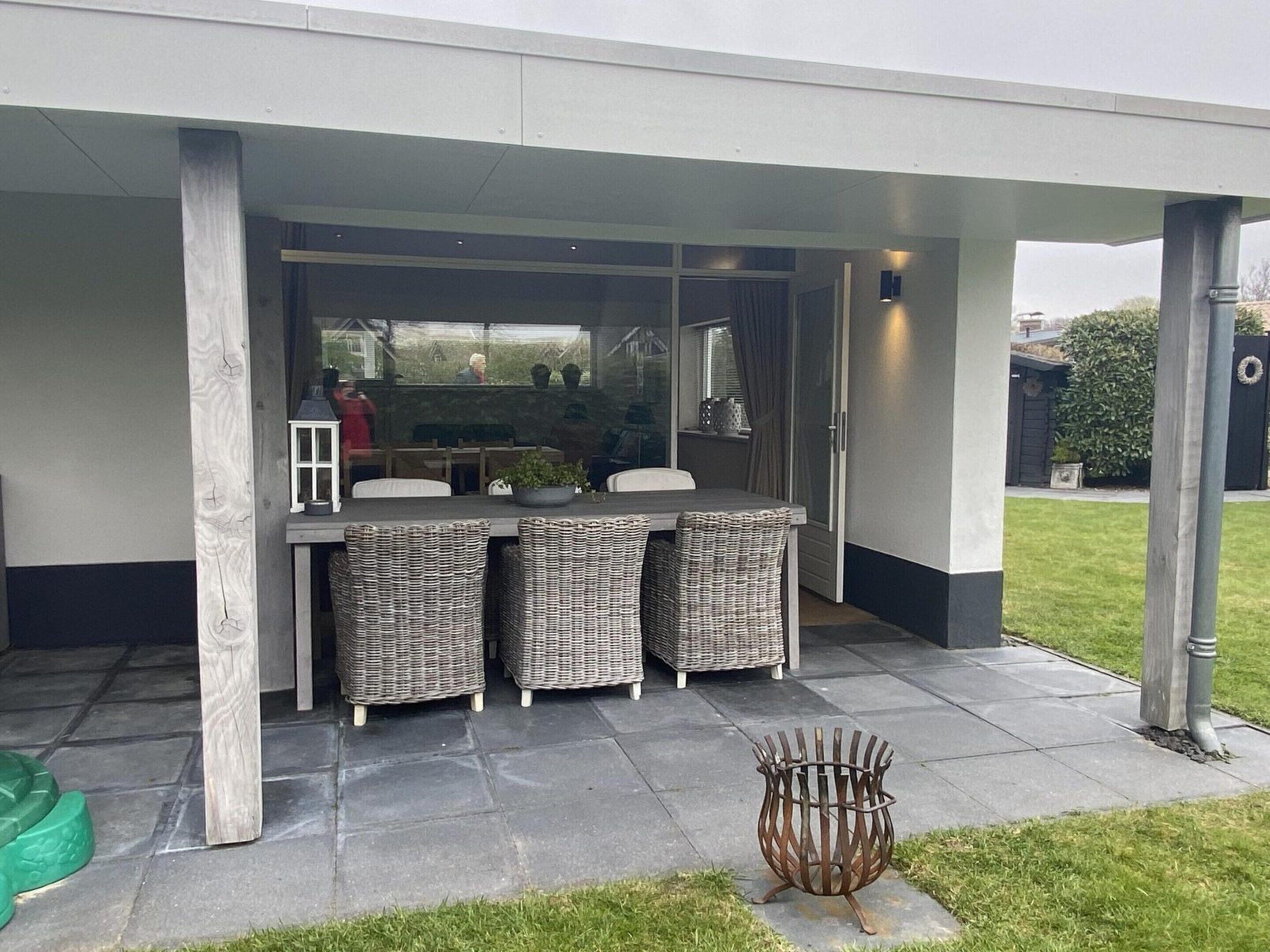 An outdoor patio with a table and wicker chairs, a metal fire pit, and a view of a house through a glass door.