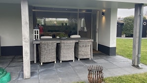 An outdoor patio with a table and wicker chairs, a metal fire pit, and a view of a house through a glass door. - Lovely Holiday Bungalow With Garden (Sint Maartensvlotbrug)