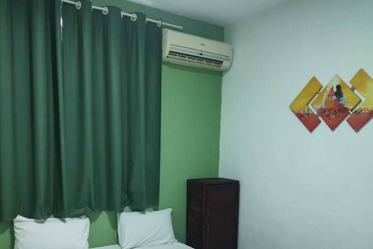 Ein Zimmer mit einem Einzelbett, einer grünen Wand, einer Klimaanlage, einem dunkelbraunen Kleiderschrank und einem an der Wand befestigten Kunstwerk.
