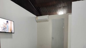 Une chambre avec un lit simple, un sol carrelé, une couvre-lit blanche, un téléviseur à écran plat fixé au mur et une porte dotée d’un miroir.