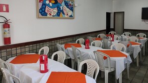 Un espace repas avec des tables recouvertes de nappes blanches et de sets de table orange, des chaises en plastique blanches, un extincteur et un tableau coloré accroché au mur.