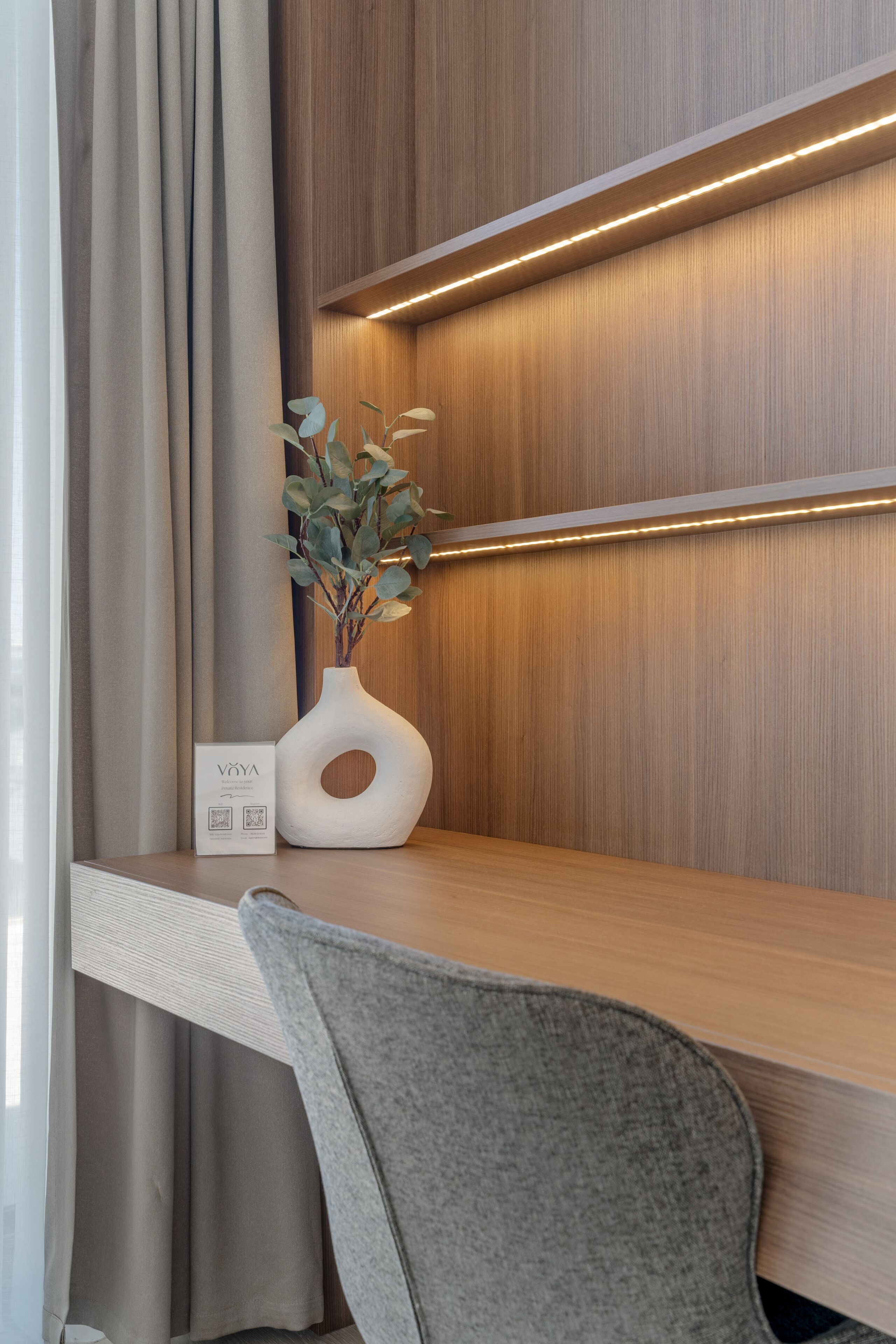 A modern hotel room with a wooden desk, a grey chair, and a vase with a plant.