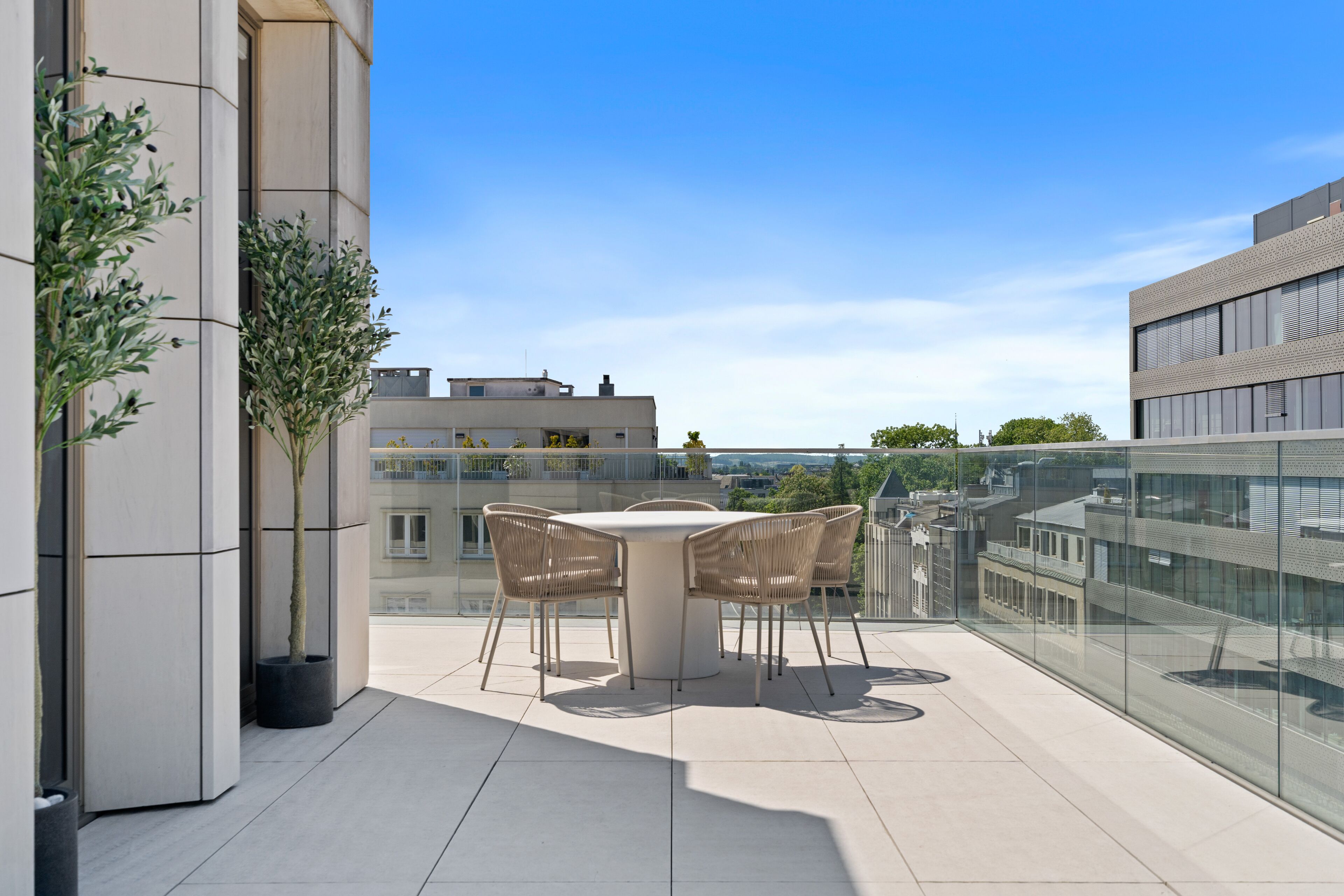 A modern outdoor terrace with a round table and chairs, potted plants, and a glass balustrade overlooking a cityscape.